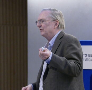 Dr. Szostak (center) gives a lecture in front of a white background
                  