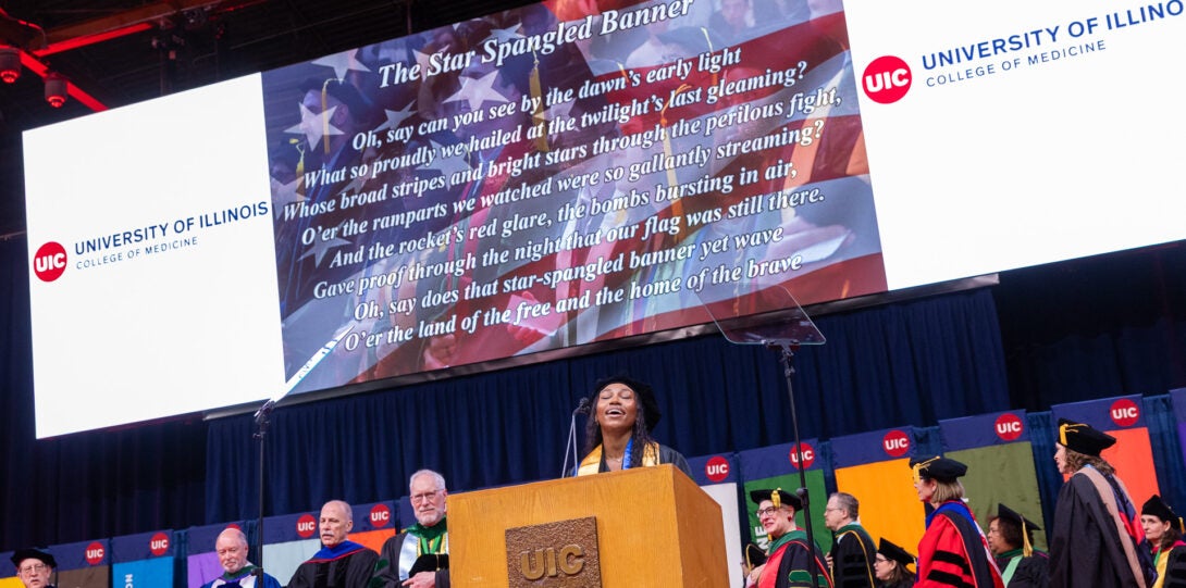 A student at the podium sings the national anthem beneath a red, white, and blue banner