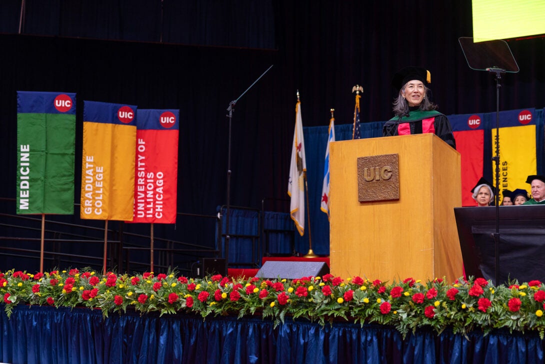 Dr. Julie Morita wears graduation gowns and speaks at the podium in front of green, yellow, and red banners.